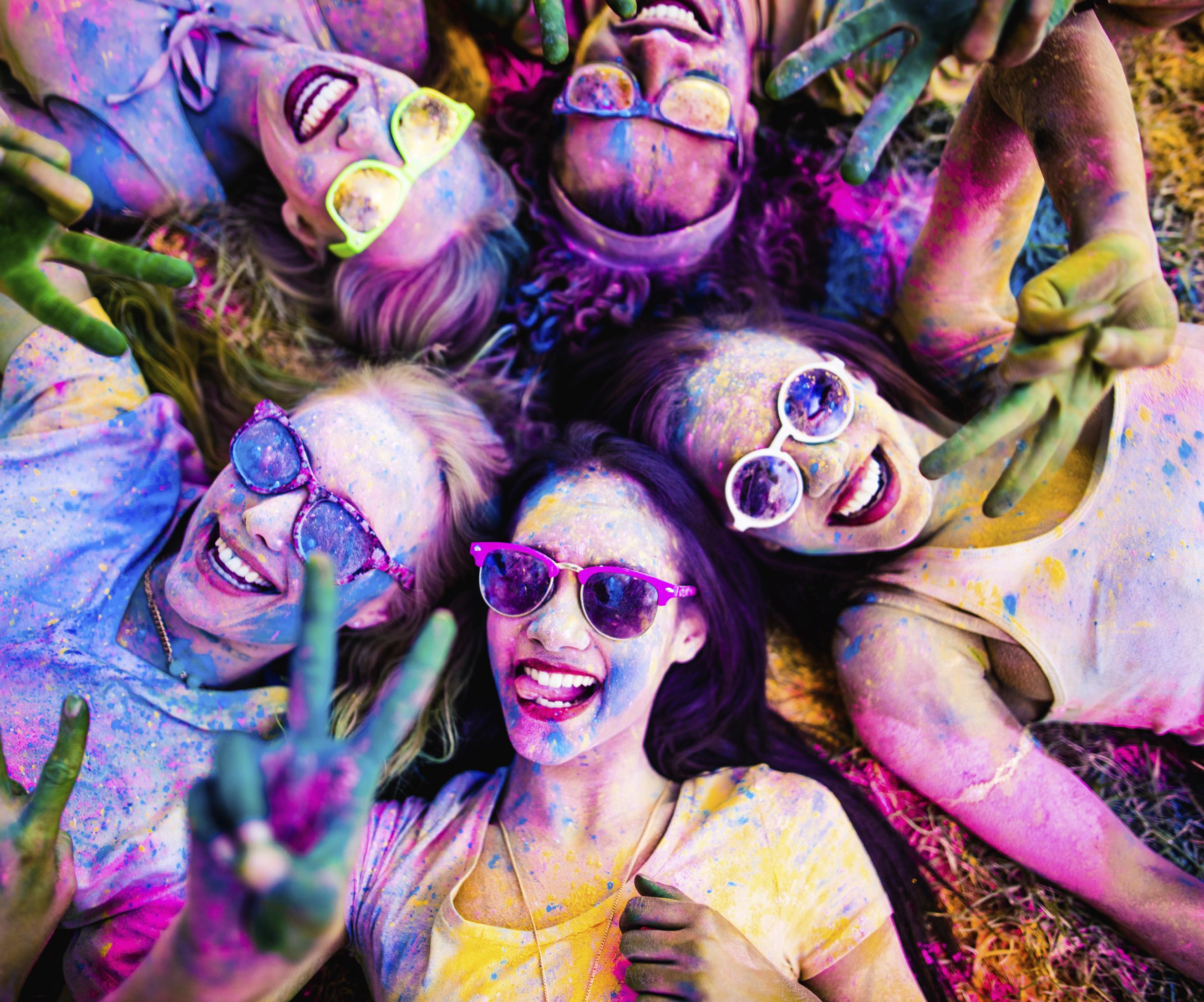 Multi-Ethnic group covered in Holi powder lay in grass laughing and looking up and gesturing peace sign to the camera in a park at a Holi festival in the summer